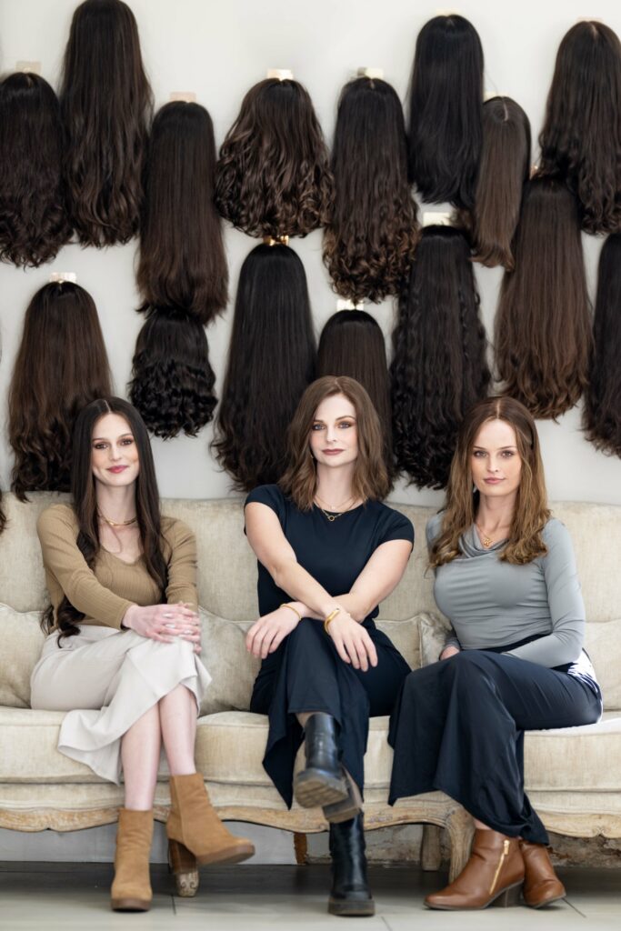 Three women sitting on couch in front of wall display of various brown and black hair wigs and extensions